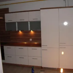 Nowoczesna zabudowa kuchenna z białymi, błyszczącymi frontami i drewnianymi akcentami, podświetlana punktowo, zrealizowana w Buk. Widoczny fragment ściany w trakcie remontu.